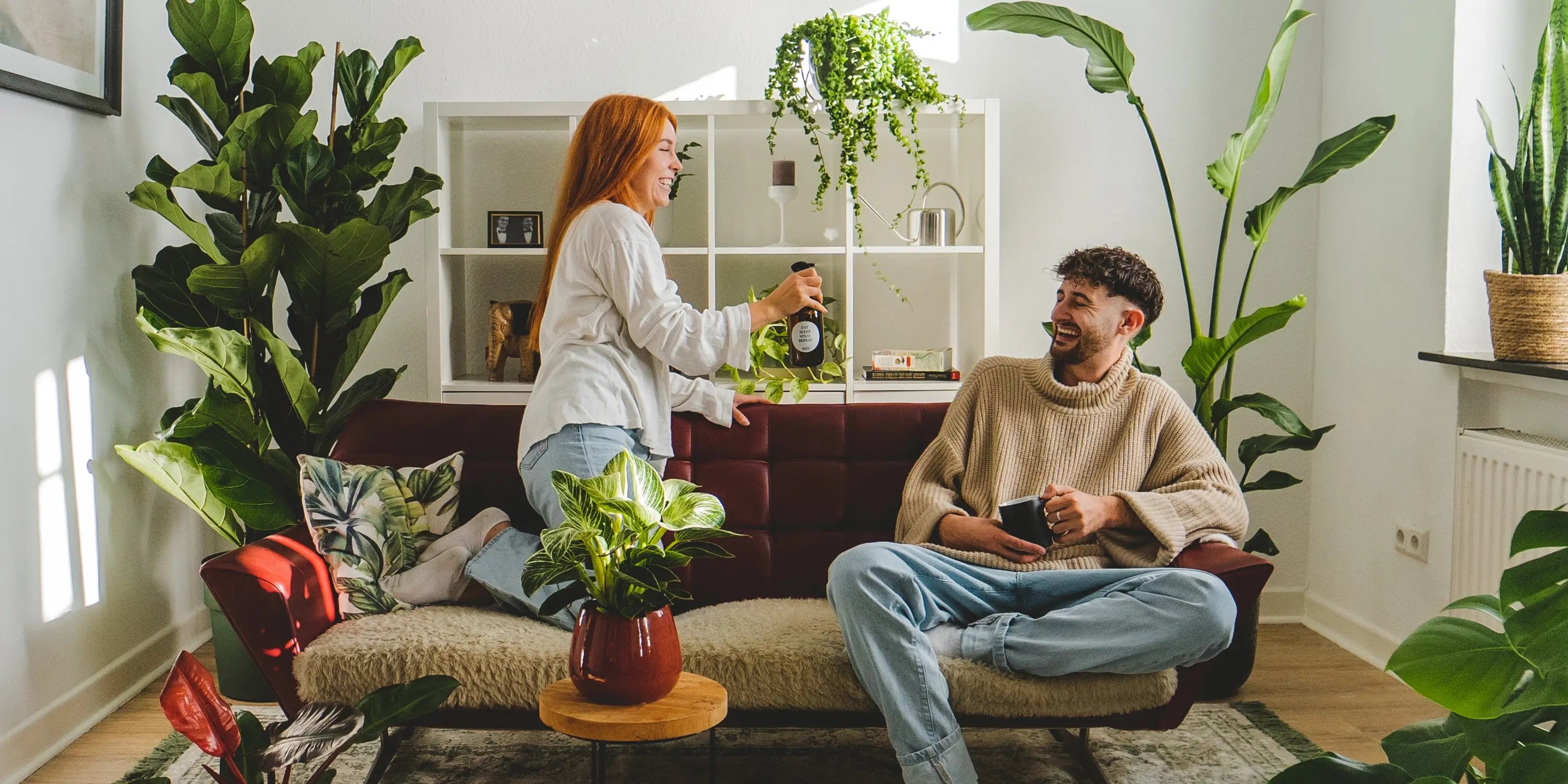 Grünes Wohnzimmer mit roter Couch voller frischer Zimmerpflanzen, Pärchen albert mit Sprühflasche herum